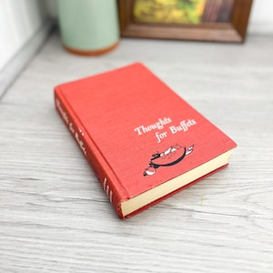 May include: A red hardcover book titled "Thoughts for Buffets" with a small illustration of a watermelon slice. The book has a textured cover and cream-colored pages. The book is angled on a light gray wooden surface.