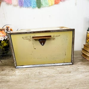May include: A rectangular metal bread box with a pale yellow finish and a weathered, distressed appearance. It features a horizontal handle and a small, dark triangular emblem centered on the front door with the text Bread and a flower icon.