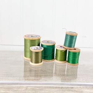 May include: A collection of six vintage wooden spools of thread in various shades of green. The spools are arranged in a group, with some stacked on top of each other. The thread is wound around the wooden spools, and the labels are visible on the top.