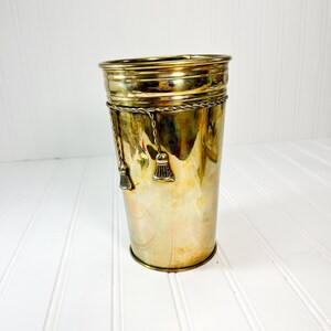 May include: A cylindrical brass umbrella stand with a shiny finish. The stand features a decorative rope-like band around the middle and two tassels hanging from the band. The stand is on a white surface.