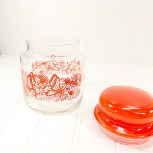 May include: A clear glass jar with a red butterfly and floral design. The jar has a matching red lid. The jar is empty and sits on a white surface.