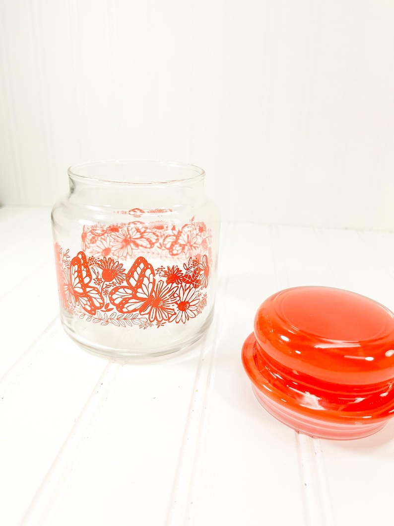 May include: A clear glass jar with a red butterfly and floral design. The jar has a matching red lid. The jar is empty and sits on a white surface.