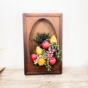 May include: Wooden wall art featuring a still life of fruit. The artwork includes red apples, yellow pears, grapes, and oranges, surrounded by green foliage. The fruit is set against a wooden background within a rectangular frame.