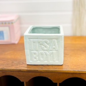 May include: A light blue ceramic planter with the words "It's a Boy!" embossed on the front. The planter is square shaped and has a smooth, glazed finish.