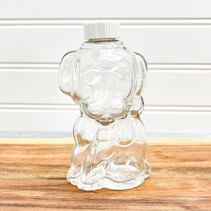 Pode incluir: Uma garrafa de vidro transparente em forma de cão com uma tampa de plástico branca. A garrafa tem detalhes em relevo e assenta numa superfície de madeira. A garrafa em forma de cão é um objeto decorativo.