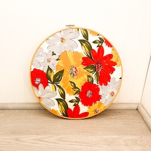 May include: A round embroidery hoop with a floral pattern. The fabric is white with red, yellow, and orange flowers.