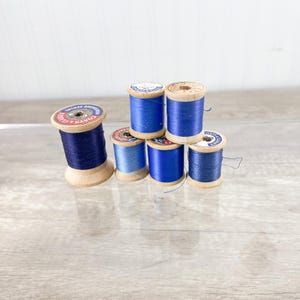 May include: A collection of vintage wooden spools of thread in various shades of blue. The spools are arranged in a small stack, with the thread wound around the wooden core. The labels on the spools are visible, adding to the vintage aesthetic.