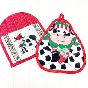May include: Two Christmas-themed kitchen pot holders. One is a mitt with a red polka dot exterior and a cow design. The other is a square pot holder with a cow design and red polka dot trim. The cow designs feature black spots, a red hat, and a green scarf.