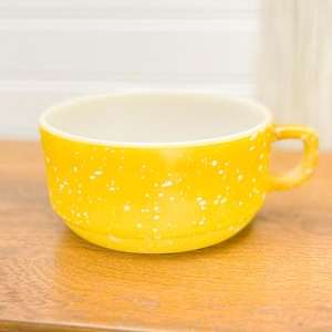 Op de afbeelding: Een gele keramische soepkom met een handvat, bespikkeld met witte stippen. De binnenkant is wit. De kom staat op een houten oppervlak. Het handvat heeft een afgeronde vorm.