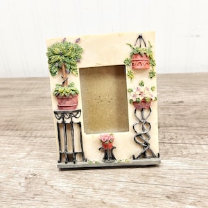 May include: Cream-colored rectangular picture frame with decorative relief of potted plants and flowers. The frame has a central opening for a photo and features a small black metal plant stand.