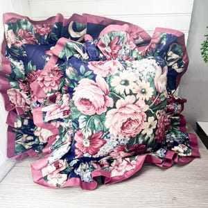 May include: A floral cushion with a frilled edge. The cushion displays a pattern of pink roses, white daisies, and green leaves on a navy blue background. The frilled edge is a solid pink colour.