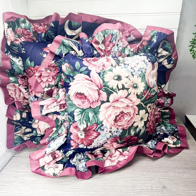 May include: A floral cushion with a frilled edge. The cushion displays a pattern of pink roses, white daisies, and green leaves on a navy blue background. The frilled edge is a solid pink colour.