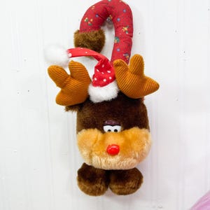 May include: A plush reindeer toy with brown fur, a tan muzzle, and a bright red nose. It features corduroy antlers and a red Santa hat with white trim. The toy is attached to a curved, red fabric handle patterned with small green and yellow shapes.