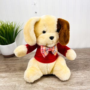 Może przedstawiać: Pluszowa zabawka w kształcie psa z kremowym futrem, brązowym uchem i czerwonym swetrem. Pies ma kraciastą muszkę i czarne oczy. Zabawka siedzi na drewnianej powierzchni.