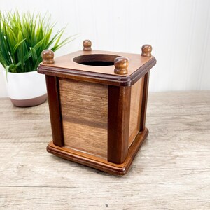 Puede incluir: Caja de pañuelos de madera de forma cuadrada y acabado marrón oscuro. La caja tiene una abertura circular en la parte superior y cuatro pequeños pomos redondeados en cada esquina. La veta de la madera es visible en la parte delantera y los laterales.