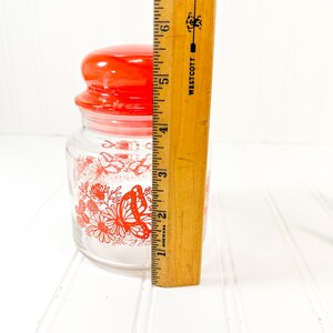 May include: A clear glass jar with a red lid and a red butterfly and floral design. The jar is approximately 4 inches tall. The jar is on a white surface.
