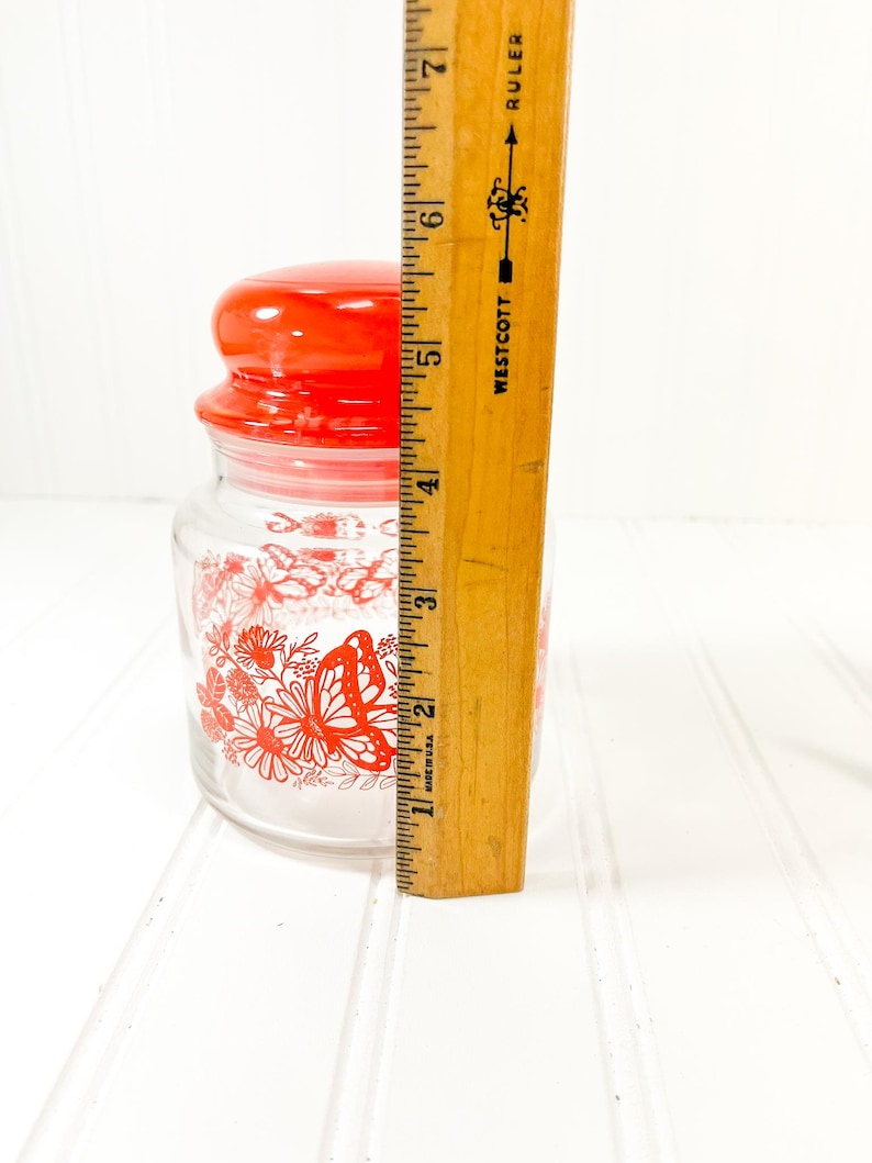 May include: A clear glass jar with a red lid and a red butterfly and floral design. The jar is approximately 4 inches tall. The jar is on a white surface.