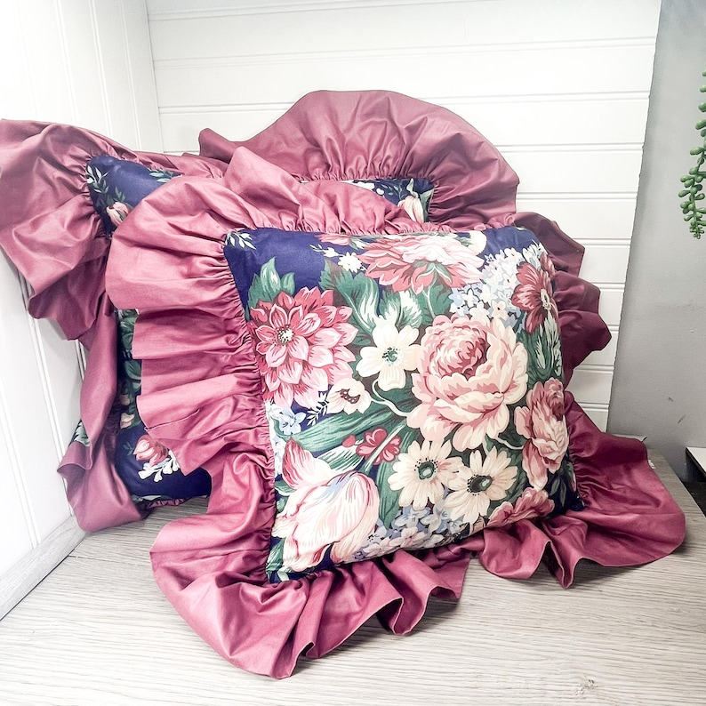 May include: Two decorative cushions with a floral pattern and frilled edges. The cushions have a dark blue background with a colourful floral design in pink, white, and green. The frilled edges are a solid mauve colour.