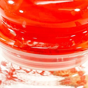 May include: A close-up of a glass jar with a bright red lid. The lid is glossy and reflects light, with a clear plastic ring beneath it. The jar's body is visible, showing a floral pattern.