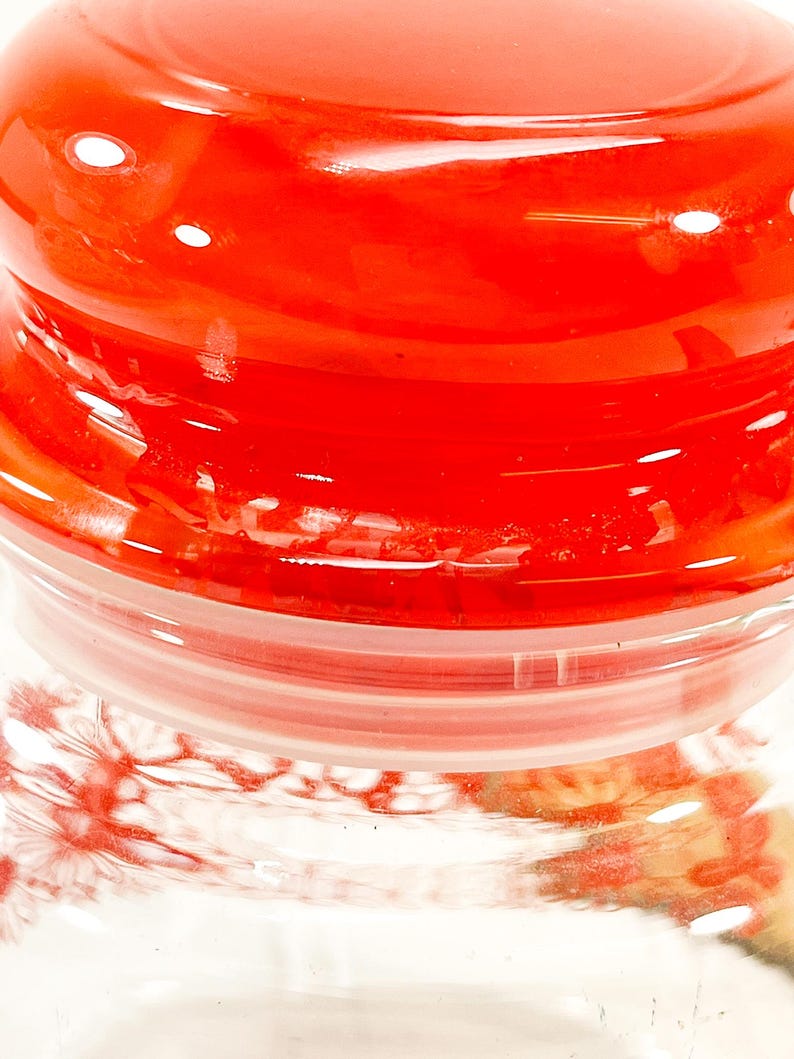May include: A close-up of a glass jar with a bright red lid. The lid is glossy and reflects light, with a clear plastic ring beneath it. The jar's body is visible, showing a floral pattern.