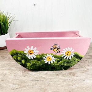 May include: A pink wooden planter with a semi-circular design. The planter is decorated with white daisies, green foliage, and a cartoon bee. The planter has two handles and is suitable for small plants.