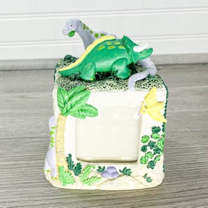 Op de afbeelding: Een decoratieve fotolijst met een groene triceratops en een grijze dinosaurus bovenop. De lijst heeft een rechthoekige opening en is versierd met een palmboom, een gele vogel en ander gebladerte. De lijst is gemaakt van licht materiaal.