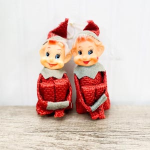 Op de afbeelding: Twee vintage kerstelf ornamenten. Elke elf heeft een lachend gezicht met blauwe ogen, een rood geweven pak en een grijze kraag. De hoed van de elf is rood met een wit touwtje om op te hangen.