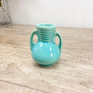 May include: A small, turquoise ceramic vase with two handles. The vase has a rounded body and a cylindrical neck with horizontal ridges. The vase is sitting on a wooden surface.