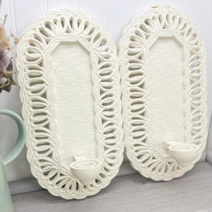 May include: Two white ceramic wall candle holders with a woven design and a scalloped edge. The holders have a simple, elegant design.