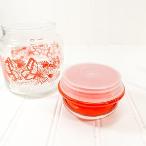 May include: Two glass jars. One is a clear jar with a red floral and butterfly design. The other is a small, round, red container with a pink lid. The jars are on a white surface.