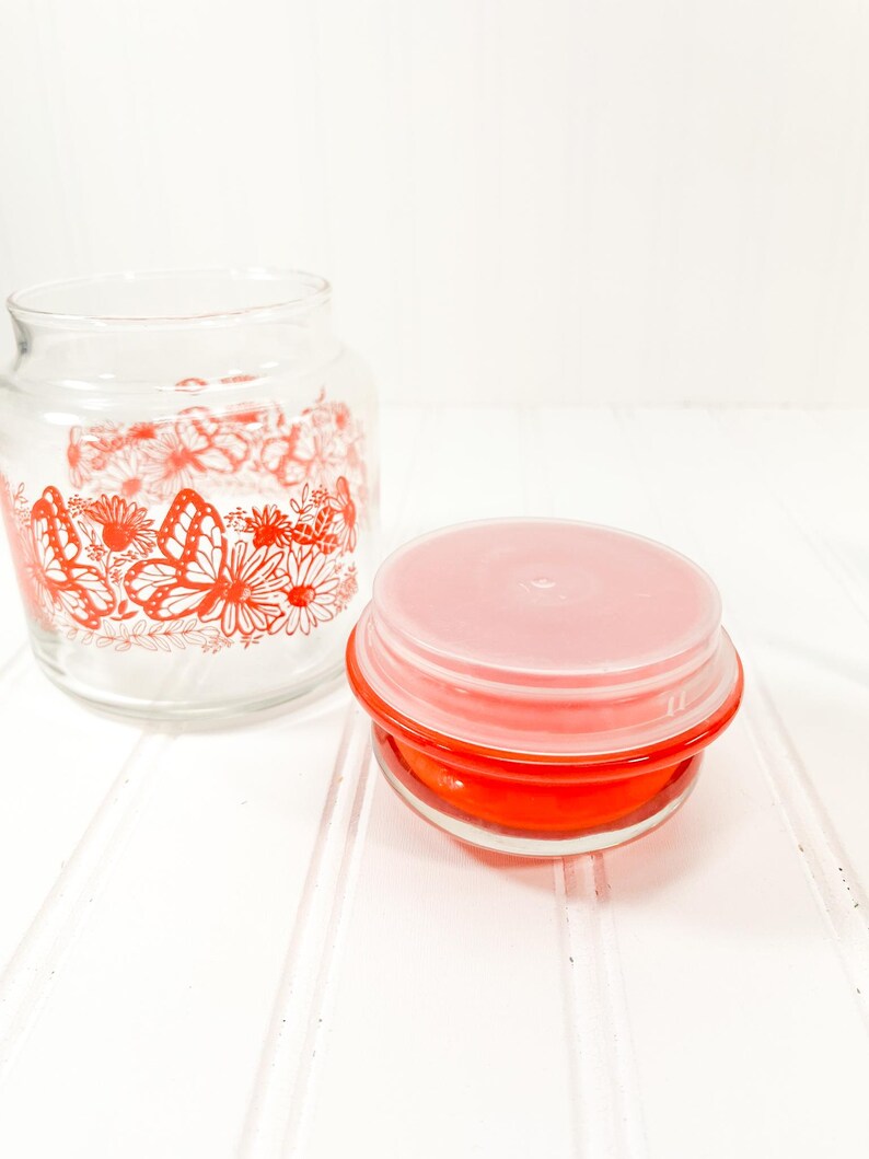 May include: Two glass jars. One is a clear jar with a red floral and butterfly design. The other is a small, round, red container with a pink lid. The jars are on a white surface.