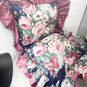 May include: Two floral patterned cushions. The cushions have a dark blue base, adorned with large pink and white flowers and green foliage. They are edged with a mauve-coloured, ruffled trim.