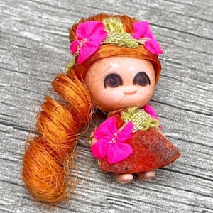 May include: A vintage doll with orange hair and a red dress. The doll has a smiling face, large brown eyes, and is adorned with pink bows and a green floral accent. The doll's hair is styled in a long ponytail.