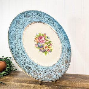 Könnte beinhalten: Vintage ovale Servierplatte mit hellblauem Rand mit einem goldenen Filigran-Design. Die Mitte hat einen Strauß bunter Blumen. Die Platte wird auf einem goldenen Metallständer präsentiert.