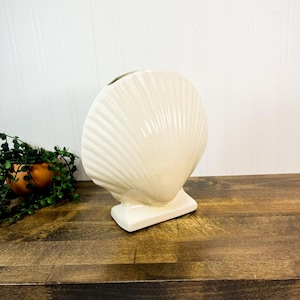 May include: Off-white ceramic seashell vase. The vase is in the shape of a scallop shell, with ribbed detailing. The vase sits on a rectangular base. The background is a wooden surface and a white wall.