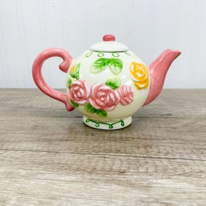以下が含まれることがあります： クリーム色の陶器製ティーポット。ピンクと黄色のバラの花と緑の葉が浮き彫りで手描きされています。ティーポットにはピンクの持ち手、ピンクの注ぎ口、そして上部に小さなピンクのつまみが付いたお揃いの蓋が付いています。