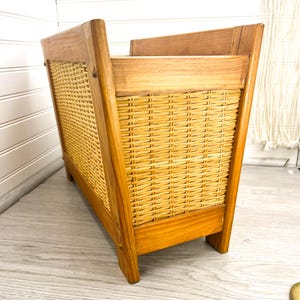 May include: A wooden magazine rack with a woven wicker front. The rack has a natural wood finish and is standing on a wooden floor.