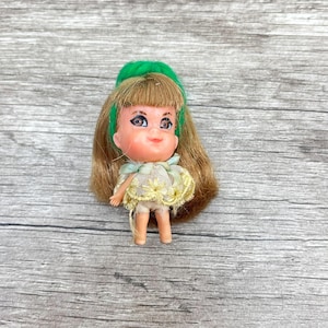 May include: A vintage doll with a green headband and long, light brown hair. The doll has a yellow and white dress with a floral pattern. The doll is small, standing approximately 3 inches tall.