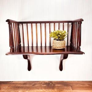 May include: A dark brown wooden wall shelf with a back and vertical spindles. The shelf has a curved front and is supported by two decorative brackets. A small potted plant in a woven basket sits on the shelf.