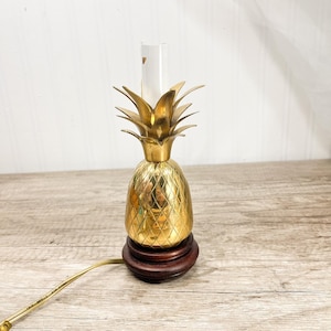 Puede incluir: Una lámpara dorada con forma de piña con una base de madera marrón. La lámpara tiene un patrón de diamantes en el cuerpo y una cubierta de vidrio transparente. La lámpara está enchufada y lista para usar.