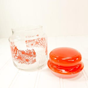 May include: A clear glass jar with a red lid, decorated with red floral and butterfly designs. The jar is open, and the lid is placed next to it. The jar is on a white surface.
