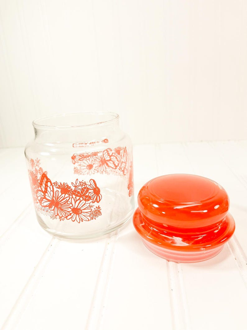 May include: A clear glass jar with a red lid, decorated with red floral and butterfly designs. The jar is open, and the lid is placed next to it. The jar is on a white surface.
