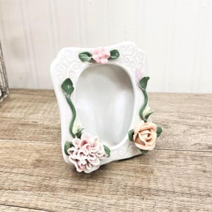 May include: White ceramic picture frame with floral accents. The frame features pink and peach-colored roses and green leaves. The frame has an oval opening and a decorative, embossed design.
