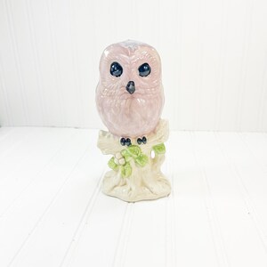 Puede incluir: Una figura de búho de cerámica en rosa suave, posada sobre un tocón de árbol color crema. El búho tiene ojos grandes y negros y una textura detallada de plumas. Hojas y bayas verdes añaden un toque de color a la base. Un artículo decorativo.