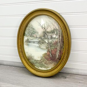 May include: Oval framed watercolor painting of a serene landscape with a river, bridge, and a white cottage. The frame is gold-colored with ornate detailing. The artwork evokes a sense of tranquility and natural beauty.