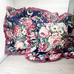 May include: Two decorative cushions with a navy blue background and a floral pattern in shades of pink, red, white, and green. The cushions have ruffled edges in a deep pink colour.