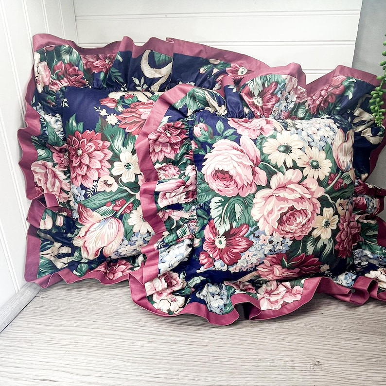 May include: Two decorative cushions with a navy blue background and a floral pattern in shades of pink, red, white, and green. The cushions have ruffled edges in a deep pink colour.