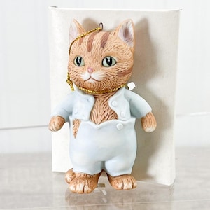 Puede incluir: Figura de cerámica de un gato con pelaje naranja y marrón, vistiendo un atuendo azul claro. El gato tiene ojos verdes y una cuerda dorada para colgar. La figura está delante de una caja blanca.