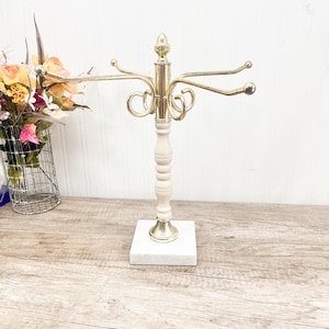 May include: A gold-toned jewelry display stand with multiple curved arms for hanging necklaces and bracelets. The stand has a white wooden center column and a square marble base. The top is adorned with a decorative finial.