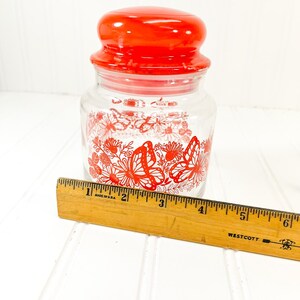 May include: A clear glass jar with a red lid and a red butterfly and floral design. The jar is approximately 4 inches tall. A wooden ruler is placed next to the jar.